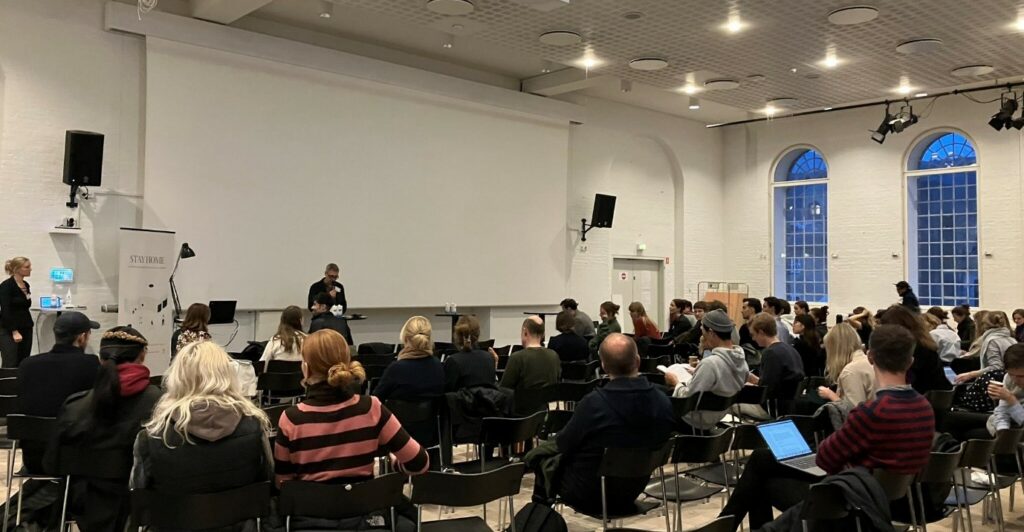 The audience of the conference sitting on chairs seen from behind. Co-PI of STAY HOME Peter Thule Kristensen introducing a keynote lecture in front of a white screen.